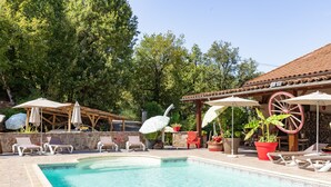Un espace piscine doté de chaises longues et de parasols, entouré d’arbres et bordé d’un bâtiment au toit recouvert de tuiles.