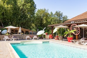 A pool area with lounge chairs and umbrellas, surrounded by trees and a building with a tiled roof. - L’Échappée Belle - joli appt avec piscine partagée (Caylus)