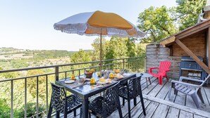 Une terrasse avec une table dressée pour les repas, des chaises d’extérieur, un barbecue et une vue sur la campagne.