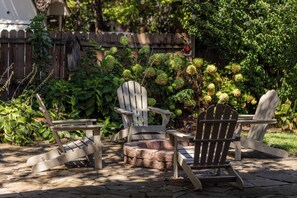 A patio area with Adirondack chairs and a stone table, surrounded by lush greenery and a wooden fence. - The Well Spoke Inn | Bike to Downtown + Coler (Bentonville)