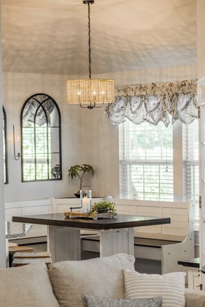 A dining area with a chandelier, a table, chairs, a window with curtains, and a plant. - The Well Spoke Inn | Bike to Downtown + Coler (Bentonville)