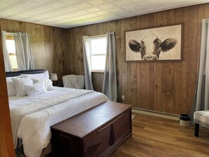 A bedroom with a wooden paneled wall, a bed with white bedding, a wooden chest of drawers, and a painting of a cow's face. - Enjoy a stay on the farm by Lake Wilhelm and Goddard State Park in Sandy Lake PA (Sandy Lake)