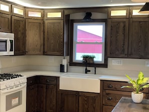 Cocina con armarios de madera, microondas, estufa, lavamanos y ventana con vista a un edificio rojo.