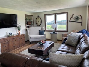 Una sala de estar con un sofá de cuero marrón, un sillón estampado, un aparador de madera, un televisor, un reloj de pared y una ventana con vistas a zonas verdes.