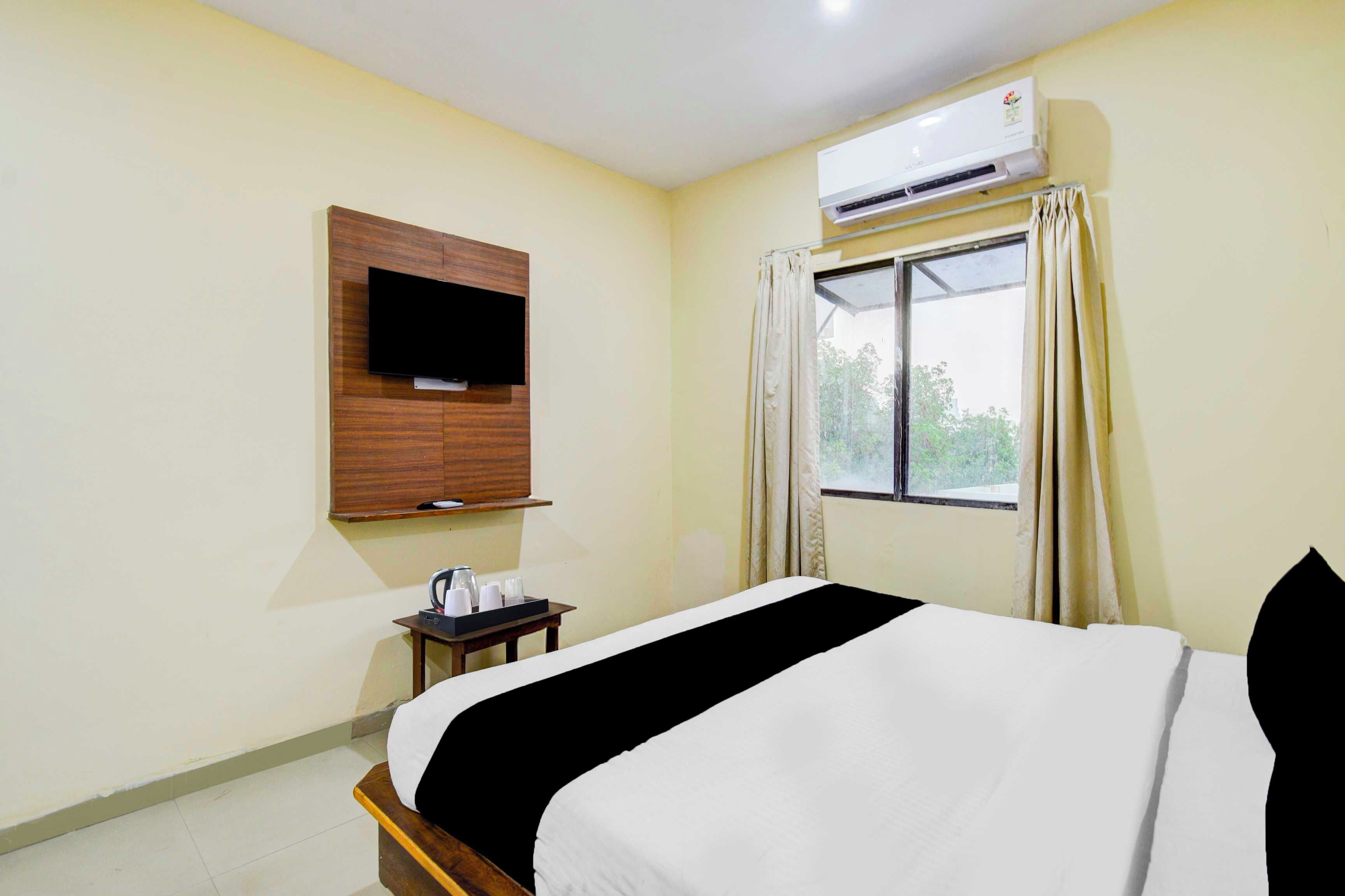 Una habitación de hotel con cama, televisión montada en la pared, equipo de aire acondicionado, ventana con cortinas y una mesita con bandeja.