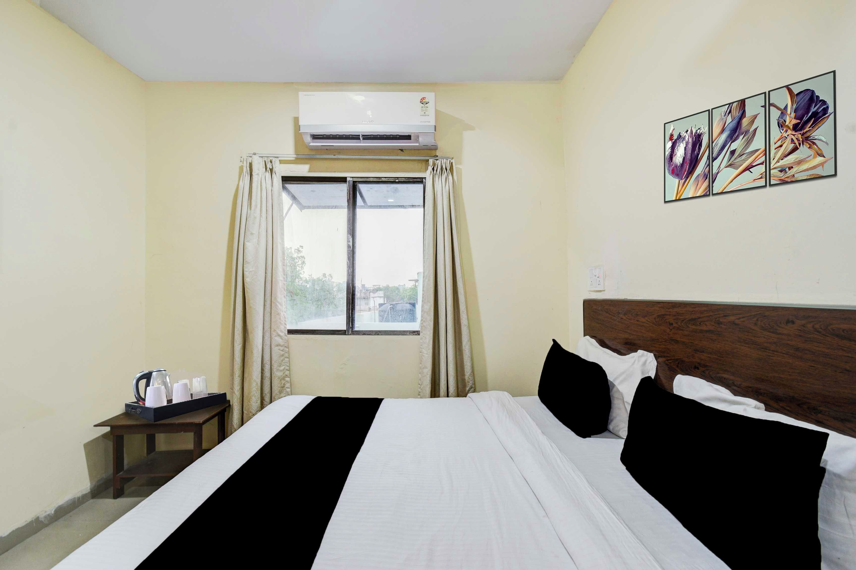 A hotel room with a bed, a bedside table with a tray of items, an air conditioning unit, a window with curtains, and a wall-mounted artwork.