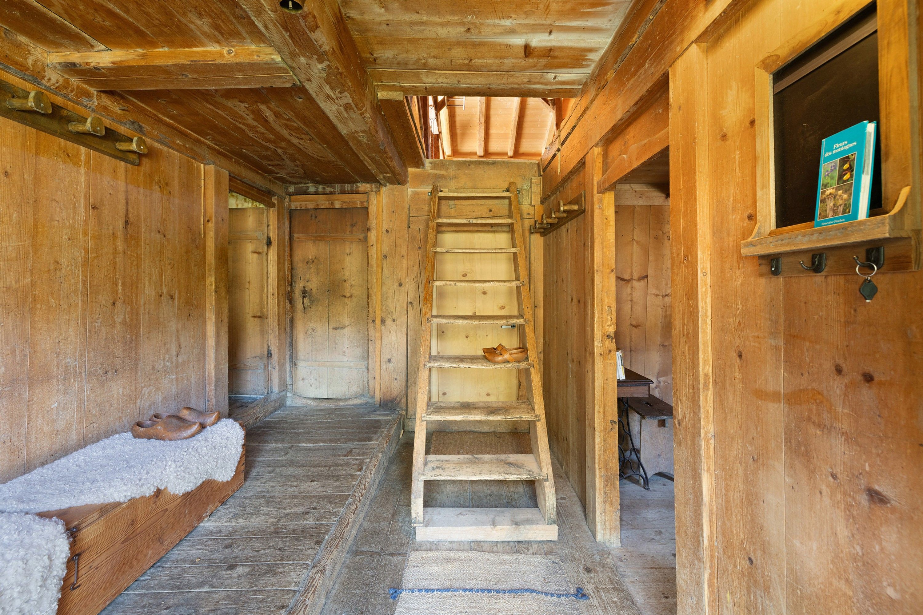 Un intérieur en bois avec un escalier central en bois menant à une mezzanine, une petite étagère avec des livres et une paire de chaussures posées sur un banc.