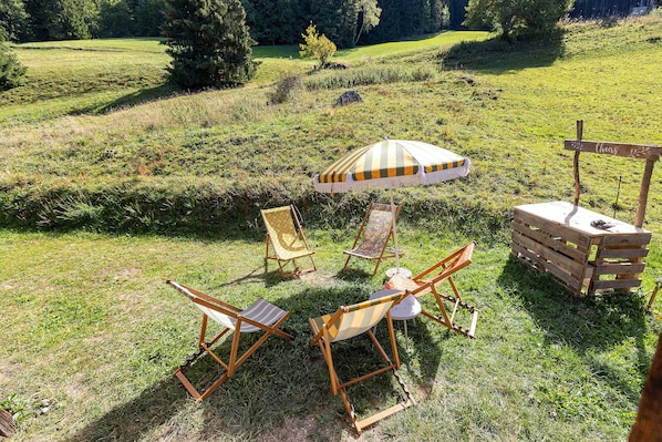 Zona salotto esterna con sedie e tavolo di legno, circondata da un prato e alberi.