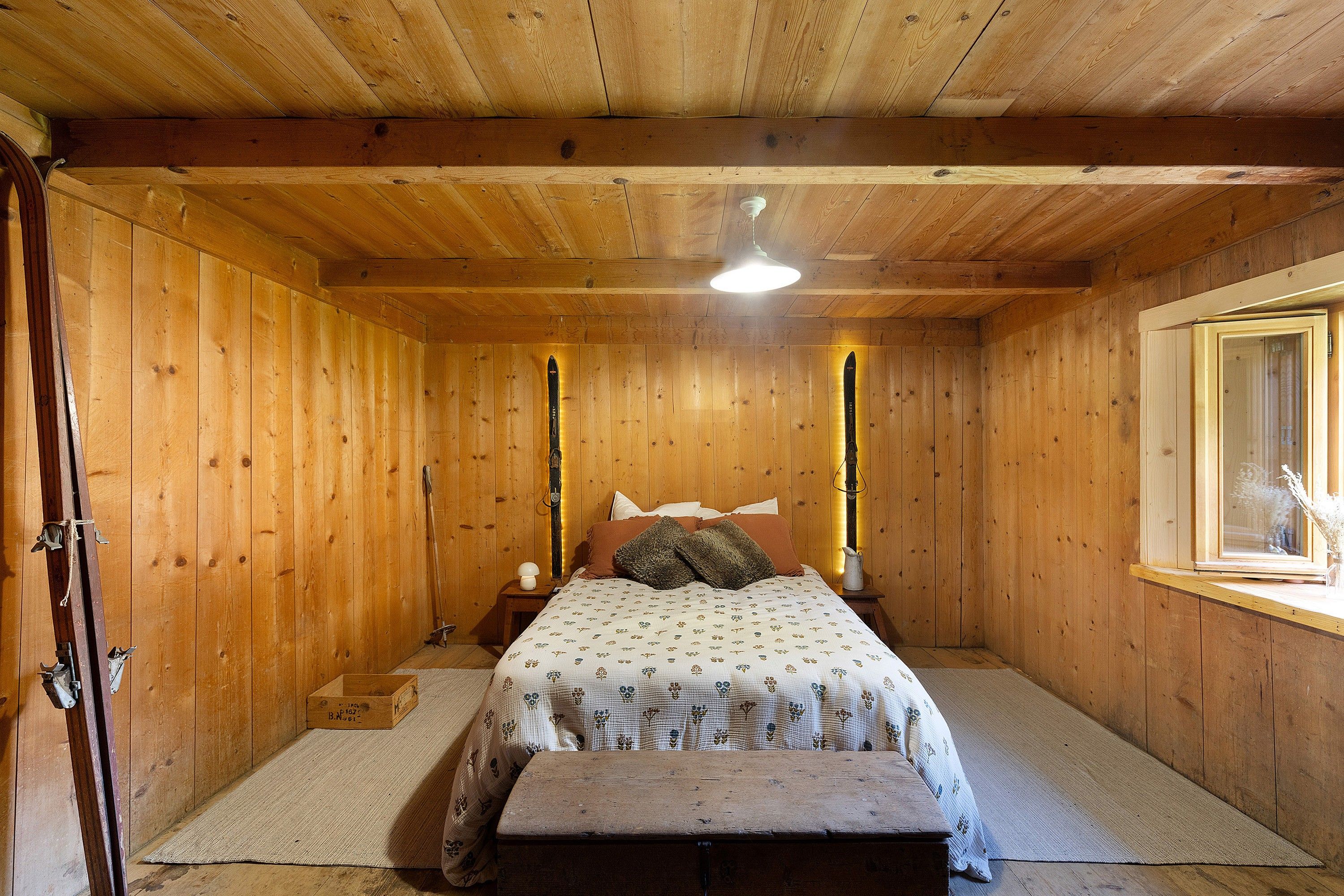 Une chambre dans une cabane en bois, avec un lit, des tables de chevet, un banc et une fenêtre.