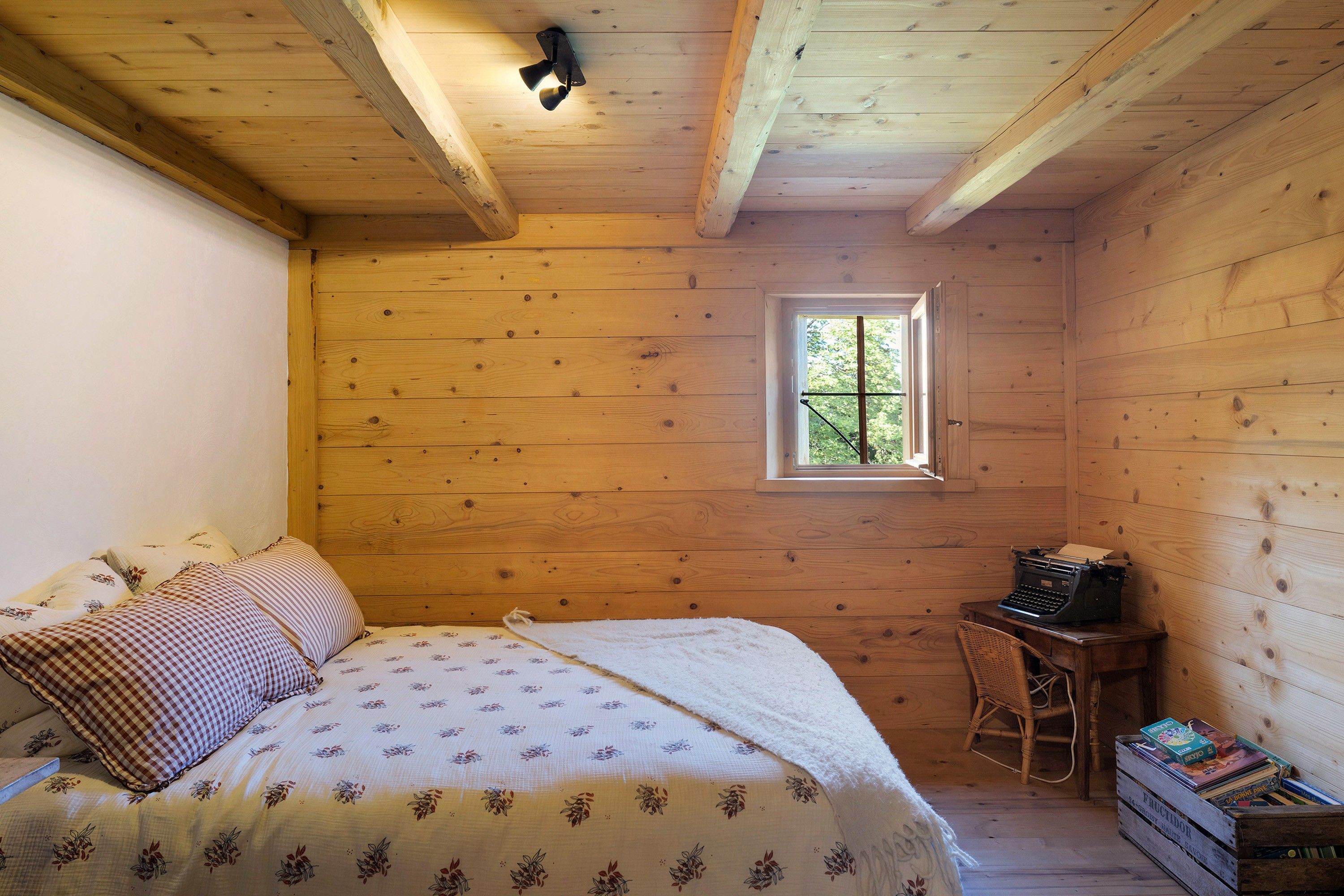 Une pièce en bois avec un lit, une petite table sur laquelle sont posés des livres, une fenêtre et un luminaire au plafond.