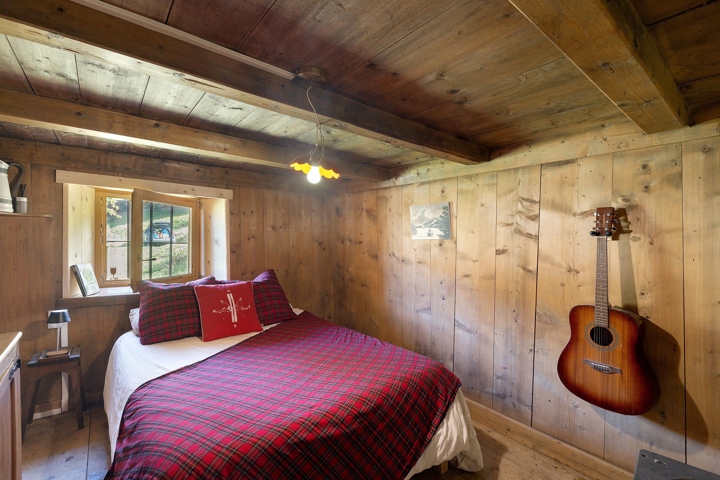 A wooden cabin room with a bed, a guitar, and a window.