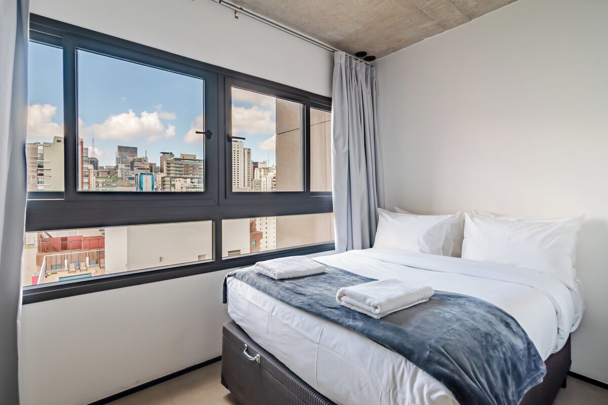 Quarto de hotel com uma cama grande, vista para a paisagem urbana através de amplas janelas e design moderno.