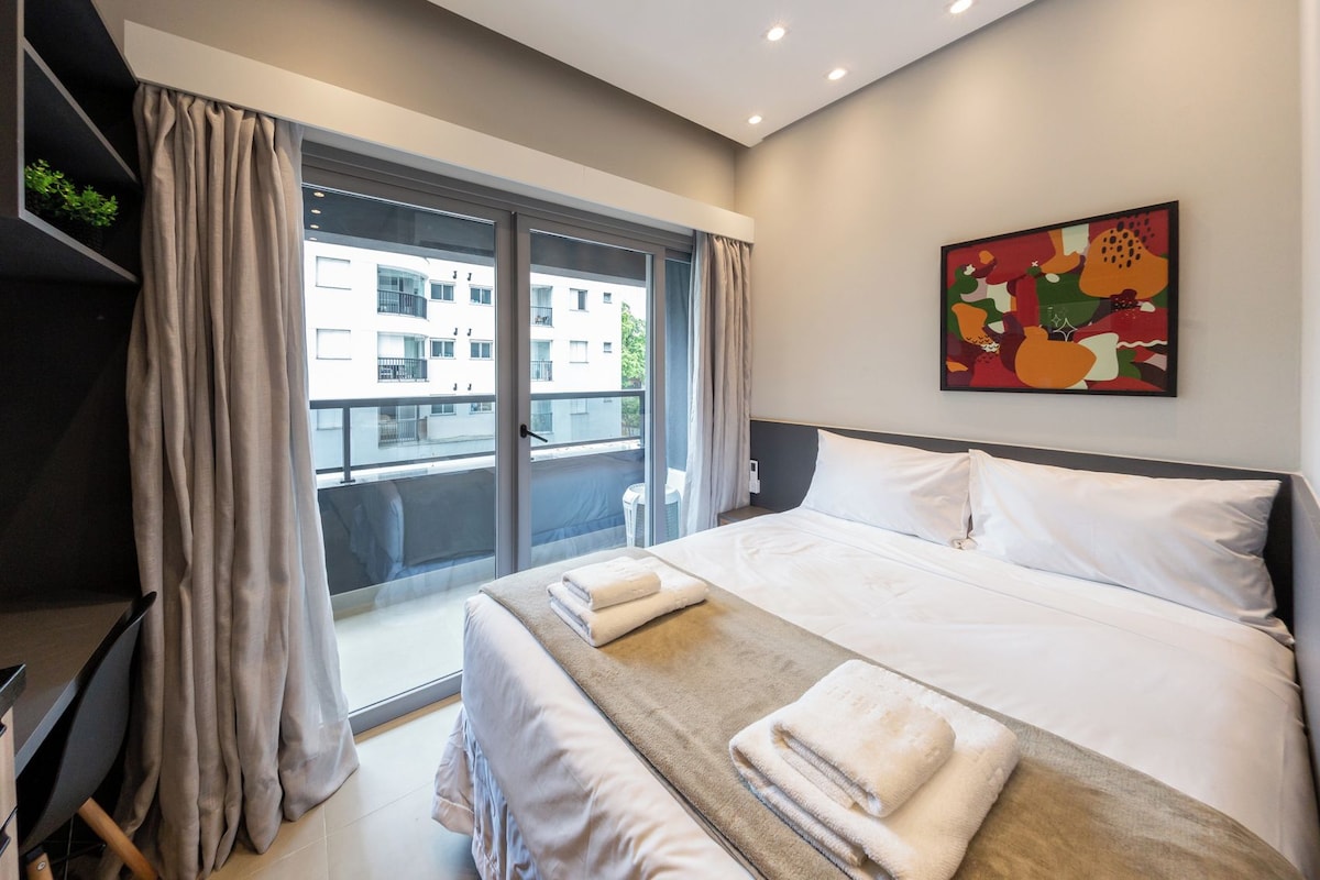 Quarto de hotel com uma cama grande, uma escrivaninha, uma cadeira, janela com cortinas e um quadro colorido na parede.