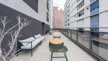 Uma área de estar moderna ao ar livre, com um sofá, cadeiras e uma mesa redonda, cercada por um parapeito de vidro.