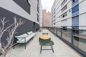 Uma área de estar moderna ao ar livre, com um sofá, cadeiras e uma mesa redonda, cercada por um parapeito de vidro.