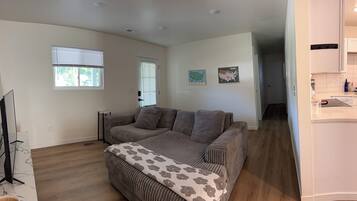 A living room with a sofa, a coffee table, and a television. There are two windows with blinds, a door, and a kitchen area visible in the background.