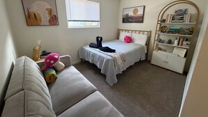 Un dormitorio con cama, mesita de noche, una silla, ventana, pared con obra de arte y estantería.