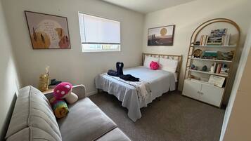 A bedroom with a bed, a bedside table, a chair, a window, a wall with artwork, and a bookshelf.