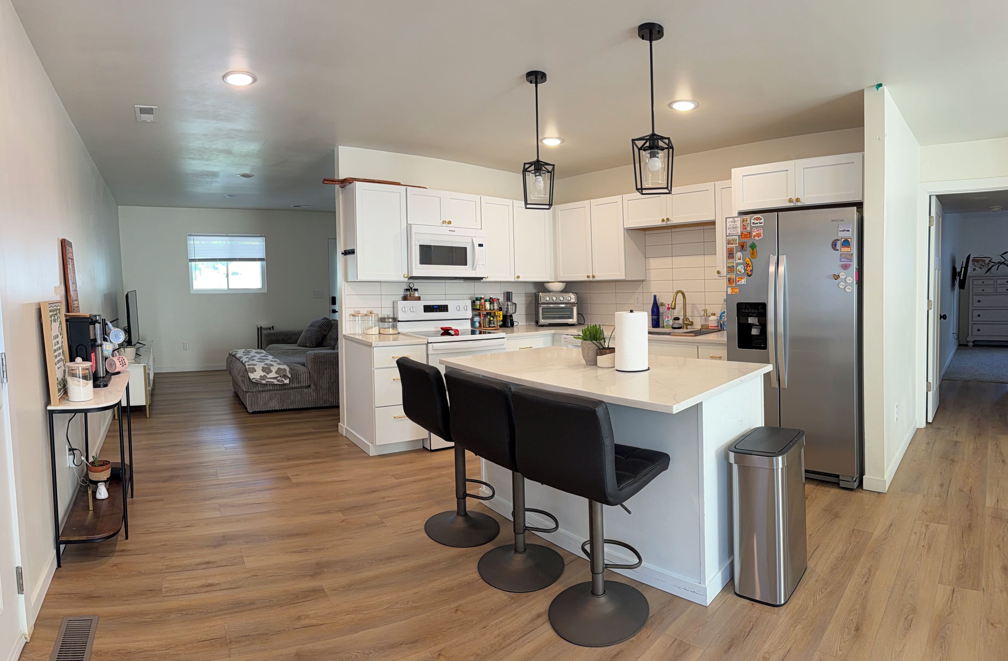 A modern kitchen with white cabinets, a stainless steel refrigerator, a microwave, and a built-in oven. There are two black bar stools and a small trash can.