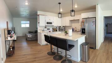 A modern kitchen with white cabinets, a stainless steel refrigerator, a microwave, and a built-in oven. There are two black bar stools and a small trash can.