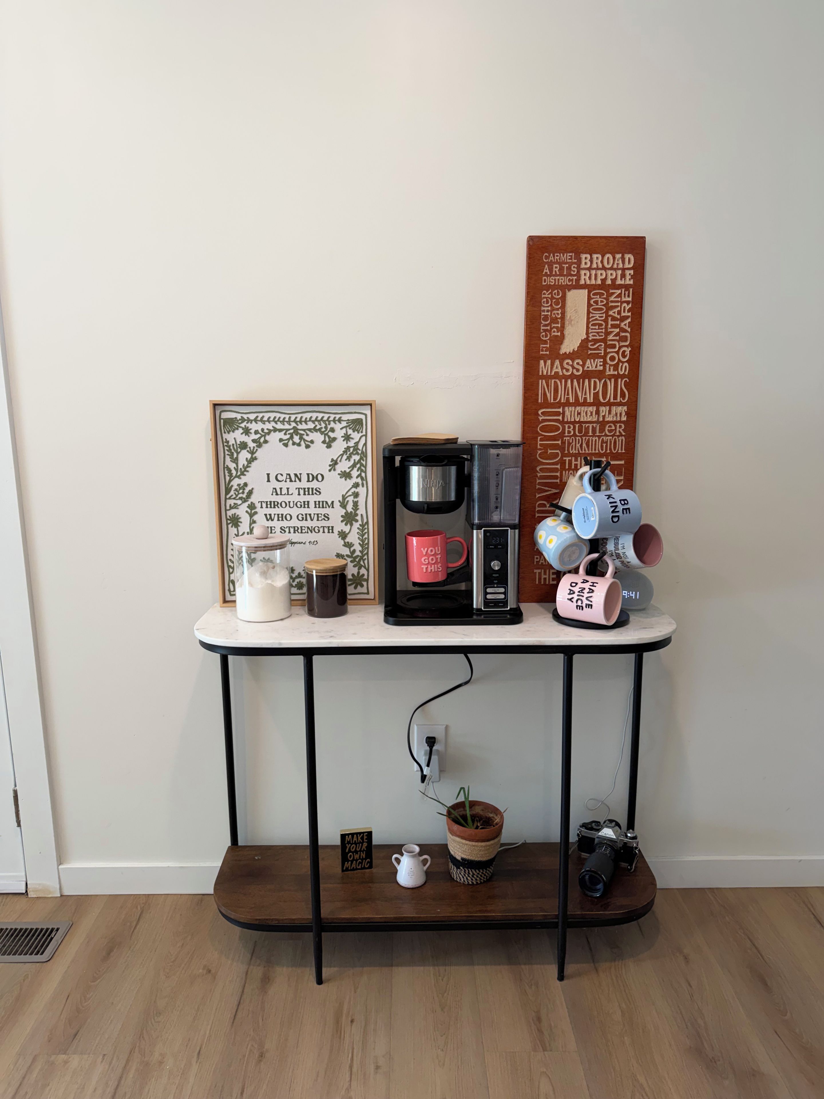 A coffee station with a coffee maker, mugs, and a motivational poster on a white table against a plain wall.
