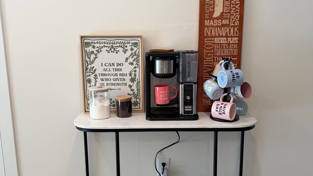 Una estación de café con cafetera, tazas y un cartel motivacional sobre una mesa blanca frente a una pared lisa.