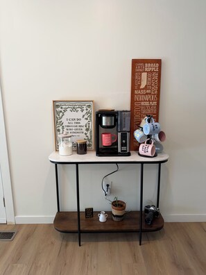 A coffee station with a coffee maker, mugs, and a motivational poster on a white table against a plain wall.