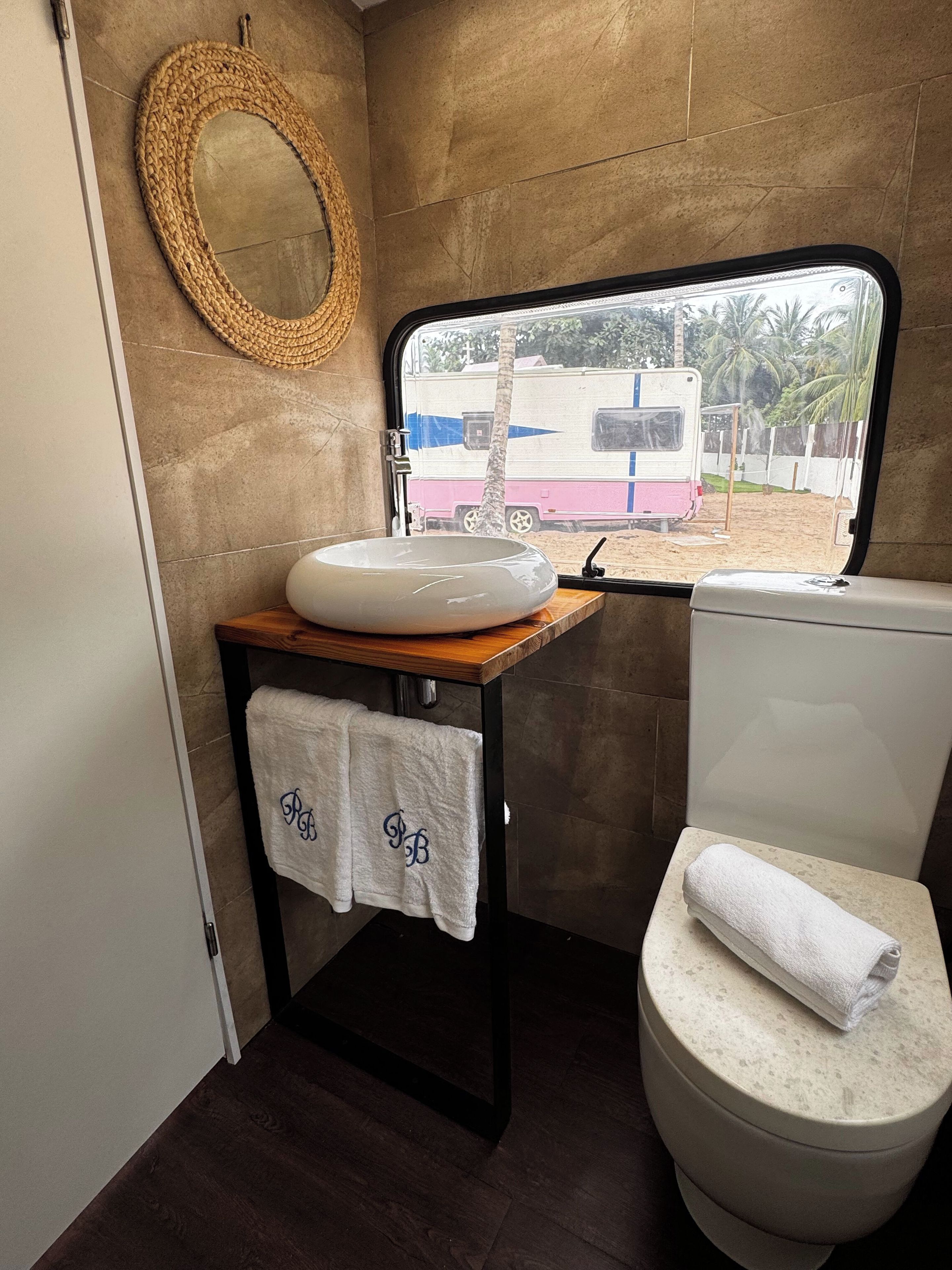 Casa de banho compacta com lavatório redondo, sanita e janela com vista para uma caravana e palmeiras.