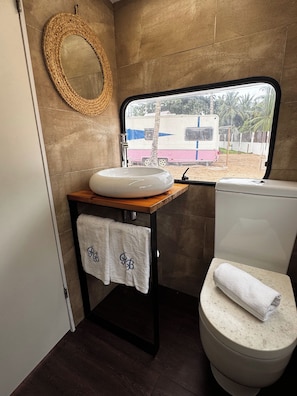 A compact bathroom with a round sink, a toilet, and a window offering a view of an RV and palm trees.
