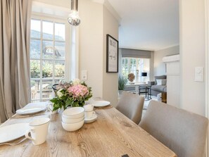 A dining area with a wooden table set for four, a vase of flowers, and a view of a house through large windows. - Holiday Home With 1 Bedroom (Sylt)