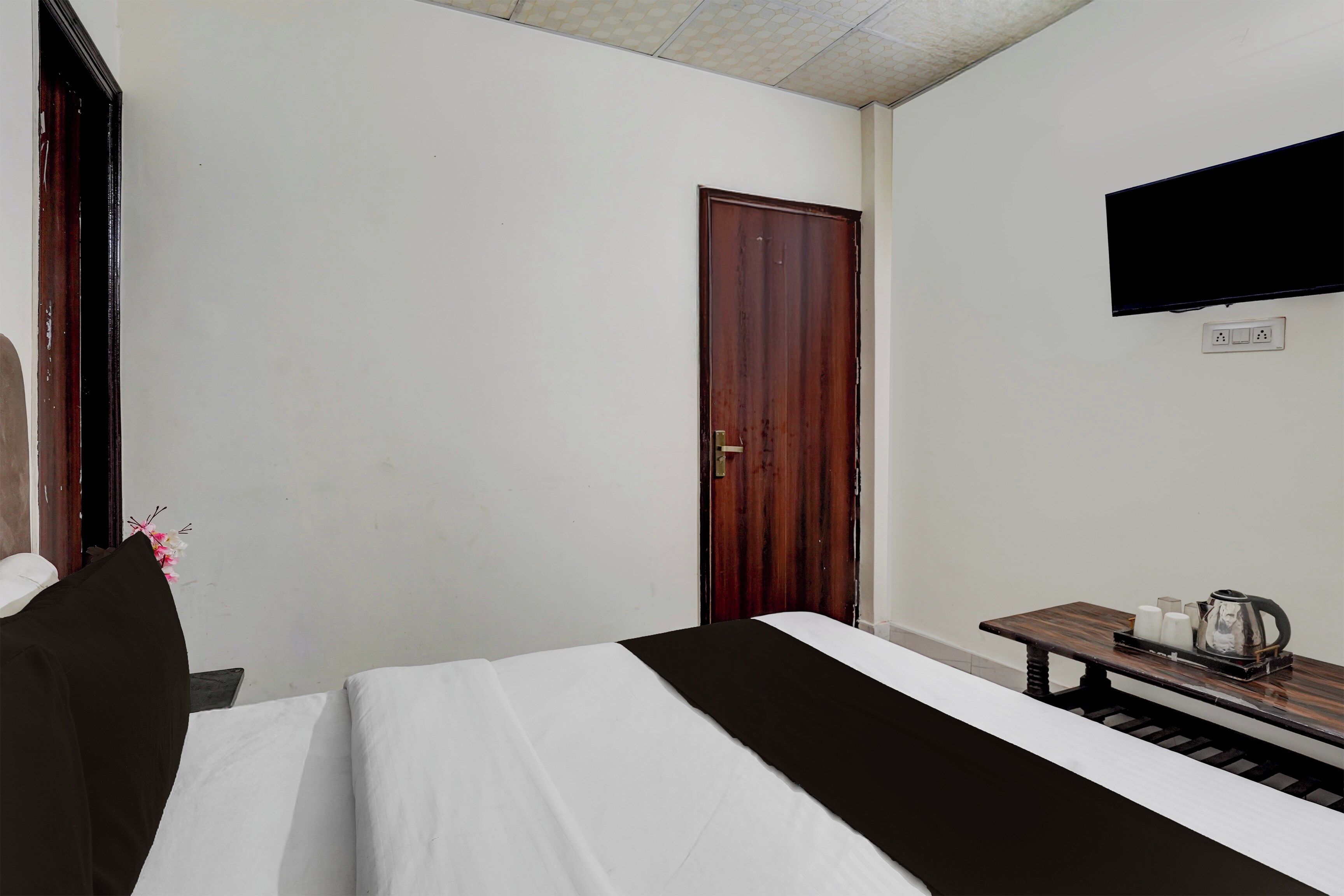 Una habitación de hotel con cama, una mesita de noche de madera con una tetera, un televisor de pantalla plana montado en la pared y una puerta.