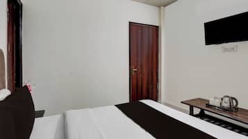 Una habitación de hotel con cama, una mesita de noche de madera con una tetera, un televisor de pantalla plana montado en la pared y una puerta.