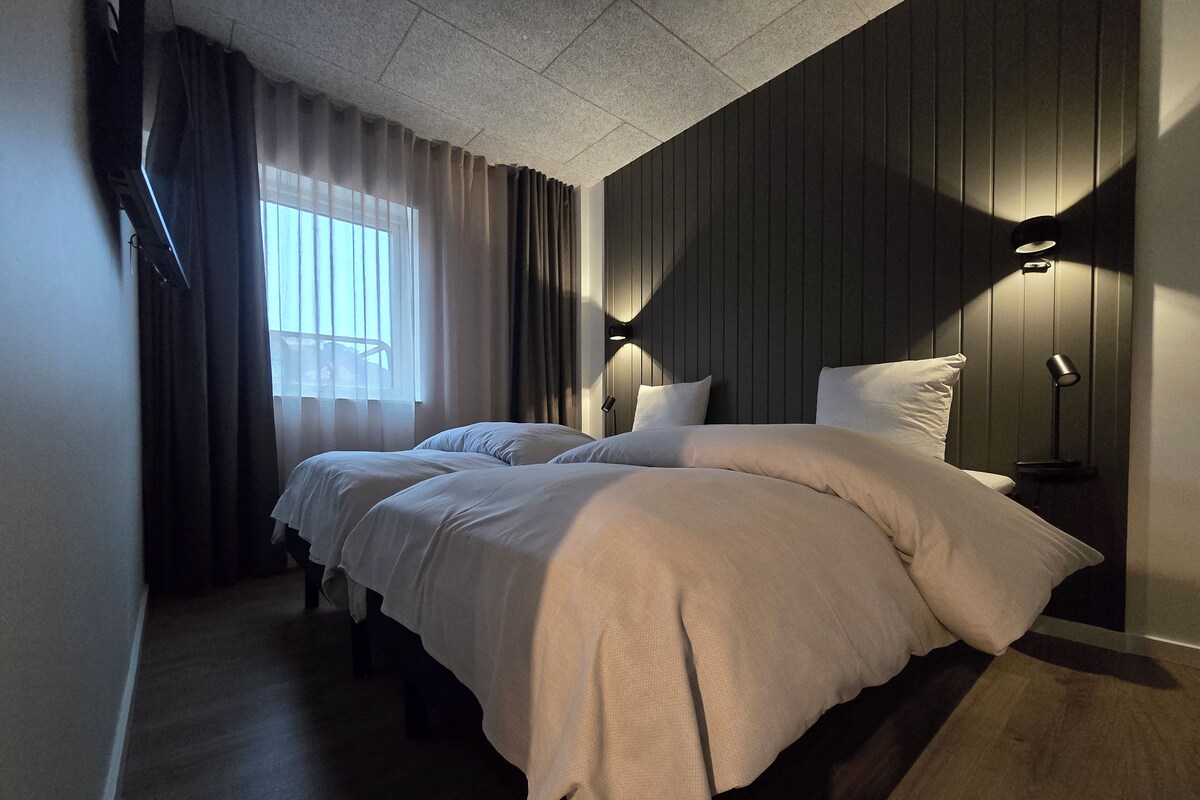 Ein Doppelbett mit Kopfteil, zwei Nachttische, ein Fenster mit Vorhängen und eine Wandlampe.
