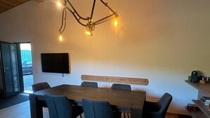 Een moderne eethoek met een grote houten tafel, zes stoelen en een aan de muur bevestigde televisie.