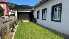 Et moderne hus med en velholdt græsplæne, en terrasse med udendørs møbler og bjergudsigt i baggrunden.