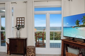 A room with a large TV displaying a beach scene, a wooden cabinet, a vase with plants, and a view of the ocean through large windows. - 🏖️Beachfront with Gulf Views | Multiple Decks | Outdoor Shower | Summer Salt🏖️ (Cape San Blas)