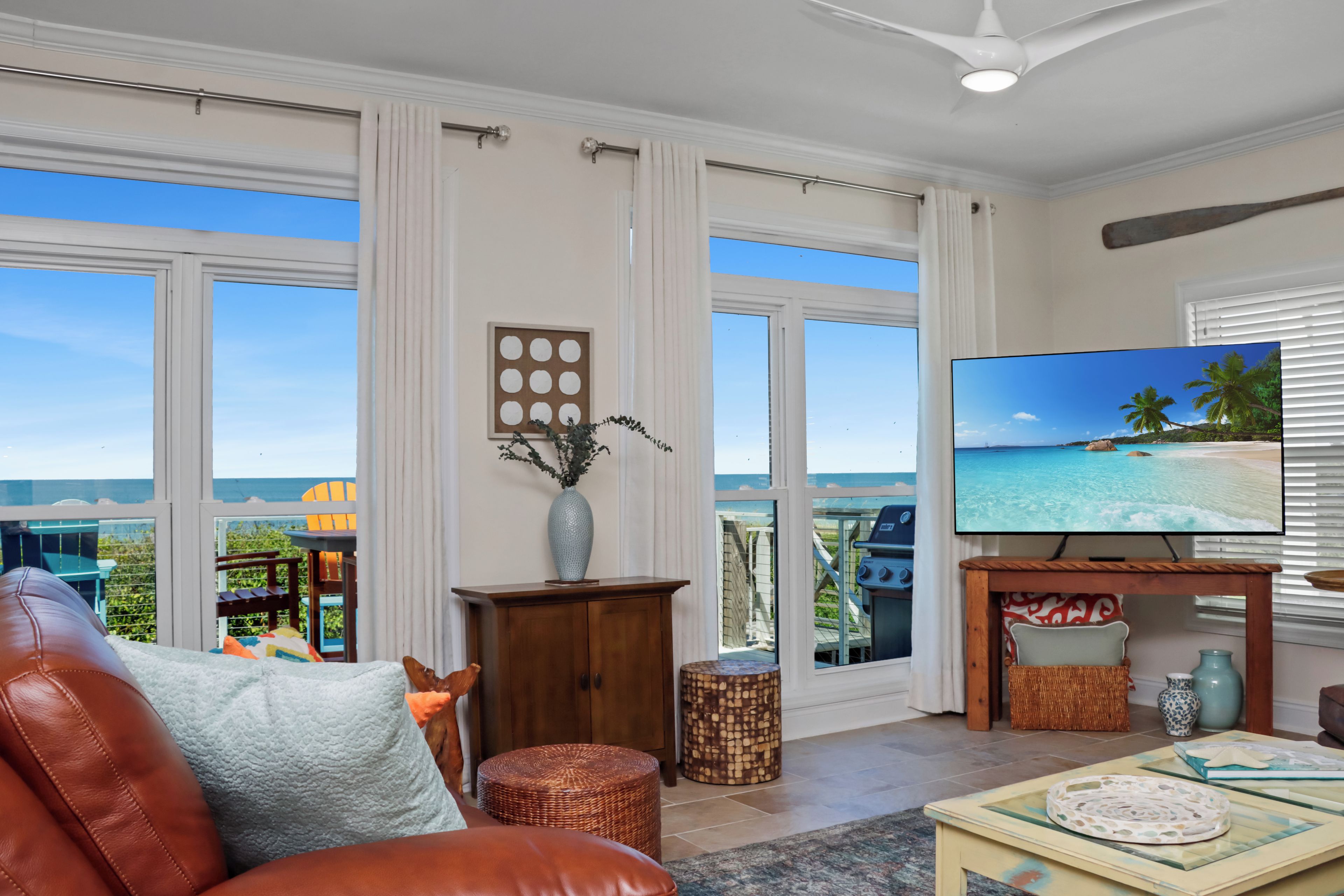 A spacious living room with a large TV, comfortable seating, and a view of the ocean through large windows.