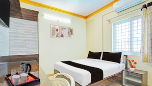 Une chambre d’hôtel comprenant un lit, une table de chevet, une fenêtre avec des rideaux, un climatiseur fixé au mur et un mur orné de trois tableaux encadrés.