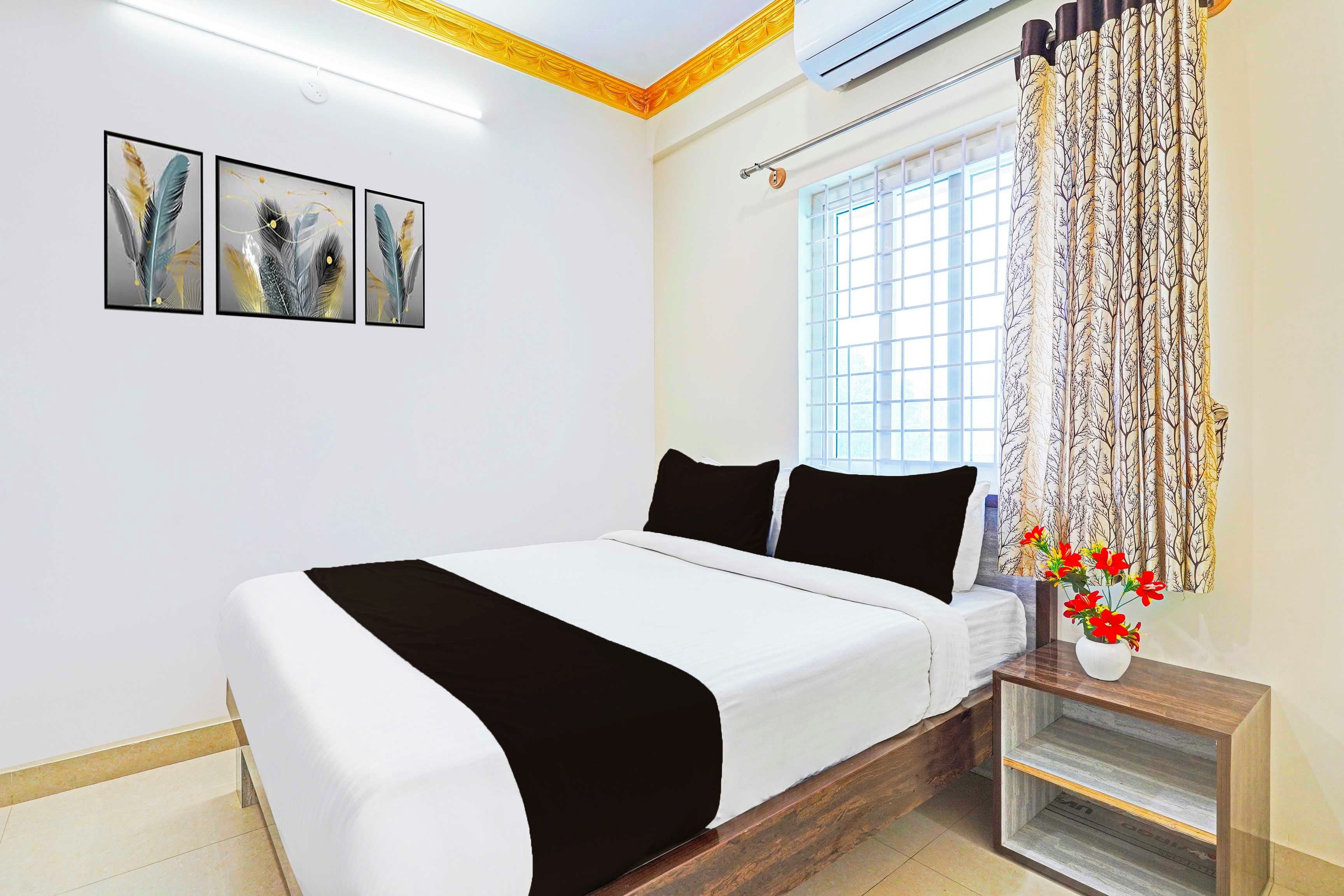 Un dormitorio con cama, ventana con cortinas, aire acondicionado de pared y un florero con flores sobre una mesita de noche.