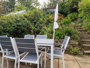 Eine Terrasse mit weißen Metallstühlen und einem Tisch, umgeben von üppiger Vegetation und einer Steinmauer.