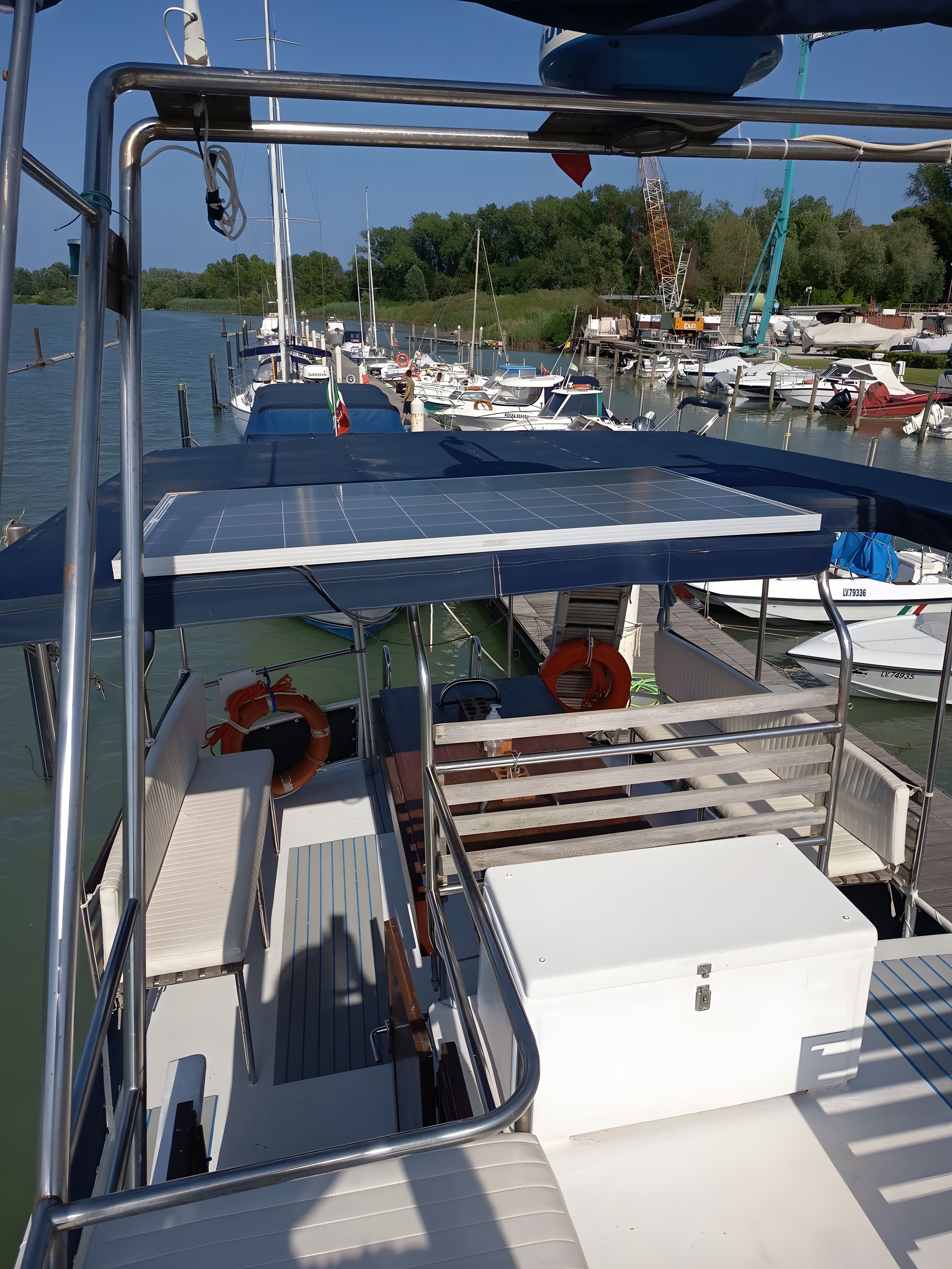 A boat docked at a marina with a covered deck, life rings, and a view of other boats and greenery in the background.