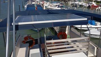 A boat docked at a marina with a covered deck, life rings, and a view of other boats and greenery in the background.