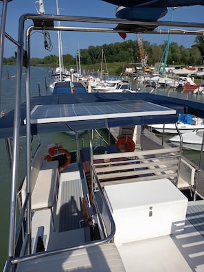 Un barco atracado en un puerto deportivo con cubierta techada, boyas salvavidas y vistas a otras embarcaciones y follaje al fondo.