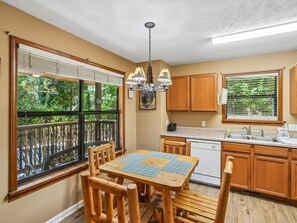 A kitchen with wooden cabinets, a dining table with chairs, a hanging light fixture, and large windows with blinds. - Bear Crest | Hot Tub, Pet-Friendly (Gatlinburg)