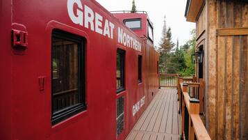 Um vagão de trem vermelho com a inscrição 'GREAT NORTHERN', um deck de madeira e vista para árvores.