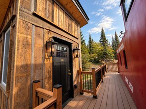 A wooden building with a black door, two wall-mounted lanterns, and a wooden deck with a railing.