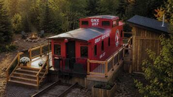 A red train caboose with 'GNX 257' and 'GREAT NORTHERN' on its side, connected to a wooden building with a porch and steps leading to a fire pit area.