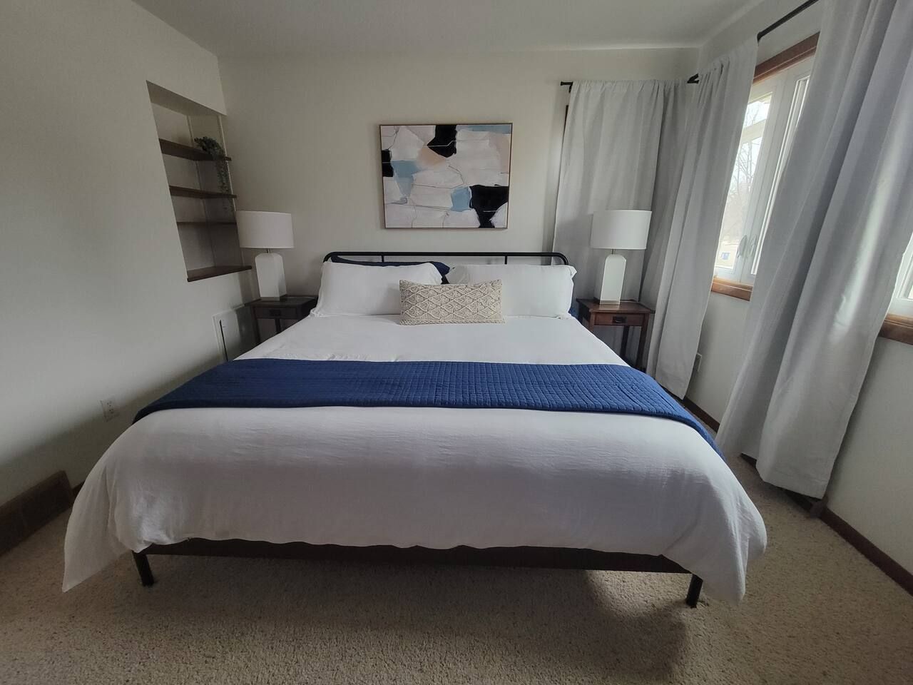 Habitació d'hotel amb un llit gran, dues taules de nit, una prestatgeria penjada a la paret i una finestra amb cortines.