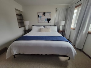 Habitació d'hotel amb un llit gran, dues taules de nit, una prestatgeria penjada a la paret i una finestra amb cortines.