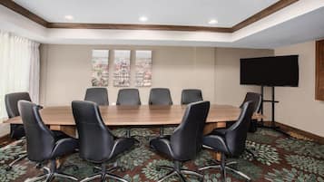 Une salle de conférence avec une longue table, des chaises noires, un téléviseur à écran plat et un tableau représentant un paysage urbain accroché au mur.